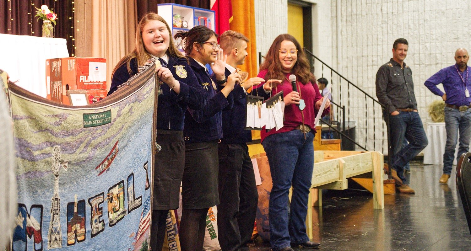 Mineola FFA members, from left, Sydney VanCleave, Valeria Veloz and Hunter Wernecke show off goods available in a non-hay lot auctioned, as emcee Jackie Rodieck describes them and Jason Ray and Don Wenecke look on. [more hay auction action]